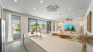 A modern kitchen with a large island, a dining area with a wooden table, and a TV mounted on the wall. - Valentine Village (Koh Samui)