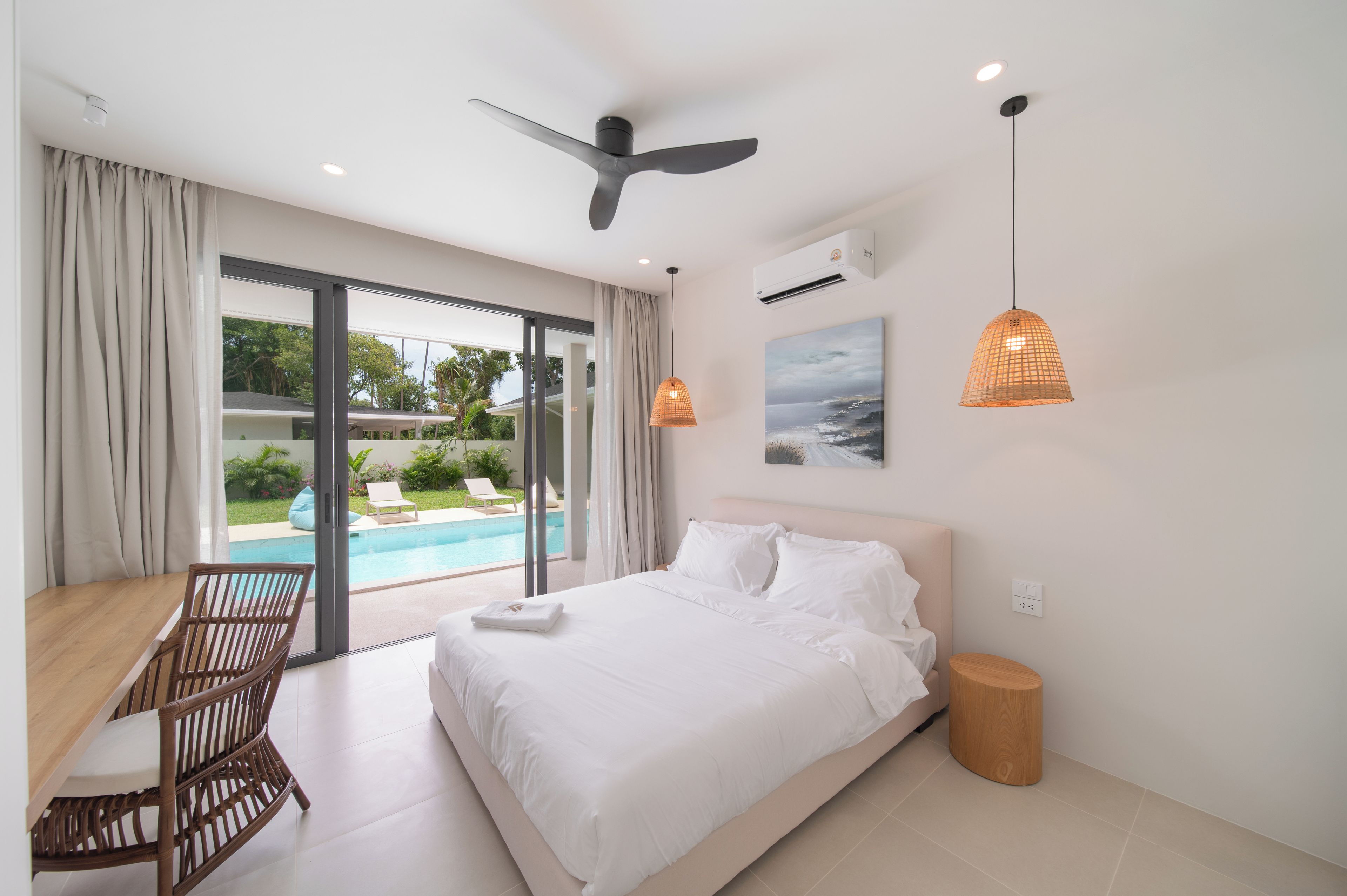 Habitación de hotel con cama, escritorio, silla, ventilador de techo, unidad de aire acondicionado, un cuadro y vista a una piscina.