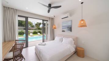 Habitación de hotel con cama, escritorio, silla, ventilador de techo, unidad de aire acondicionado, un cuadro y vista a una piscina.