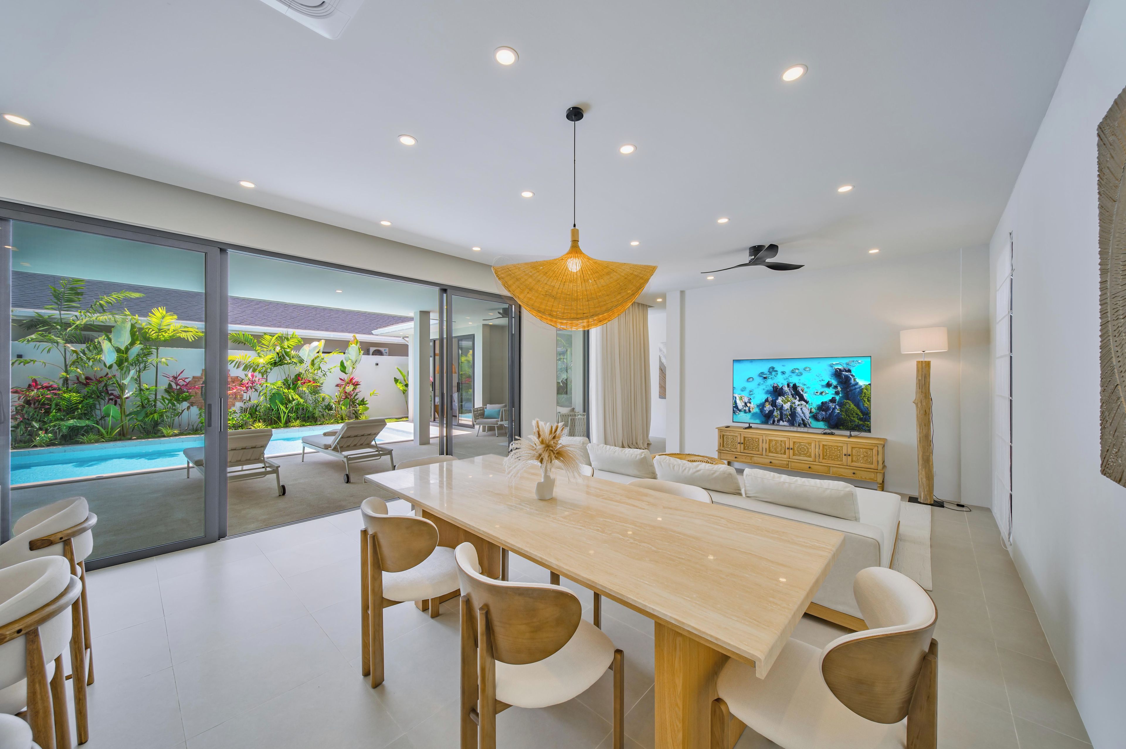A modern dining area with a wooden table and chairs, a television, and a view of a swimming pool and greenery.