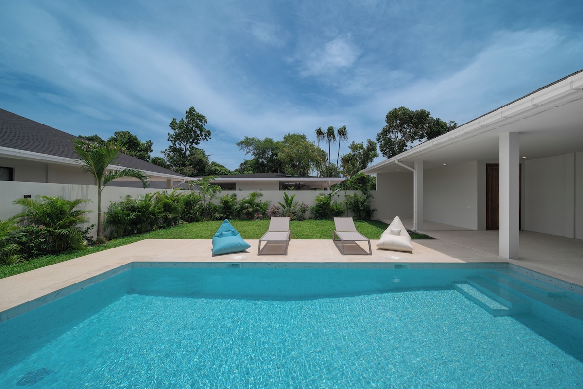 A modern pool area with clear blue water, two lounge chairs, and a small garden with various plants.