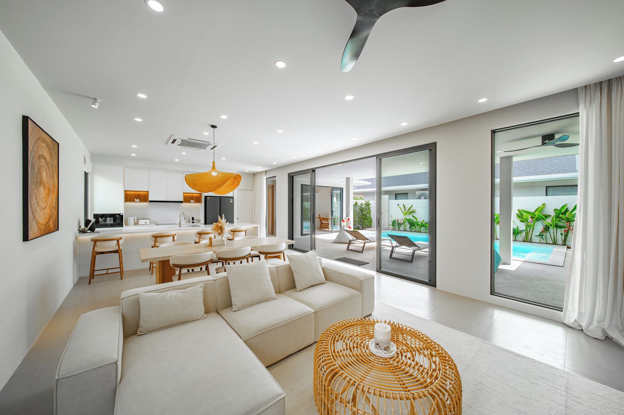 A modern living room with a sofa, dining area, and large windows overlooking a pool.