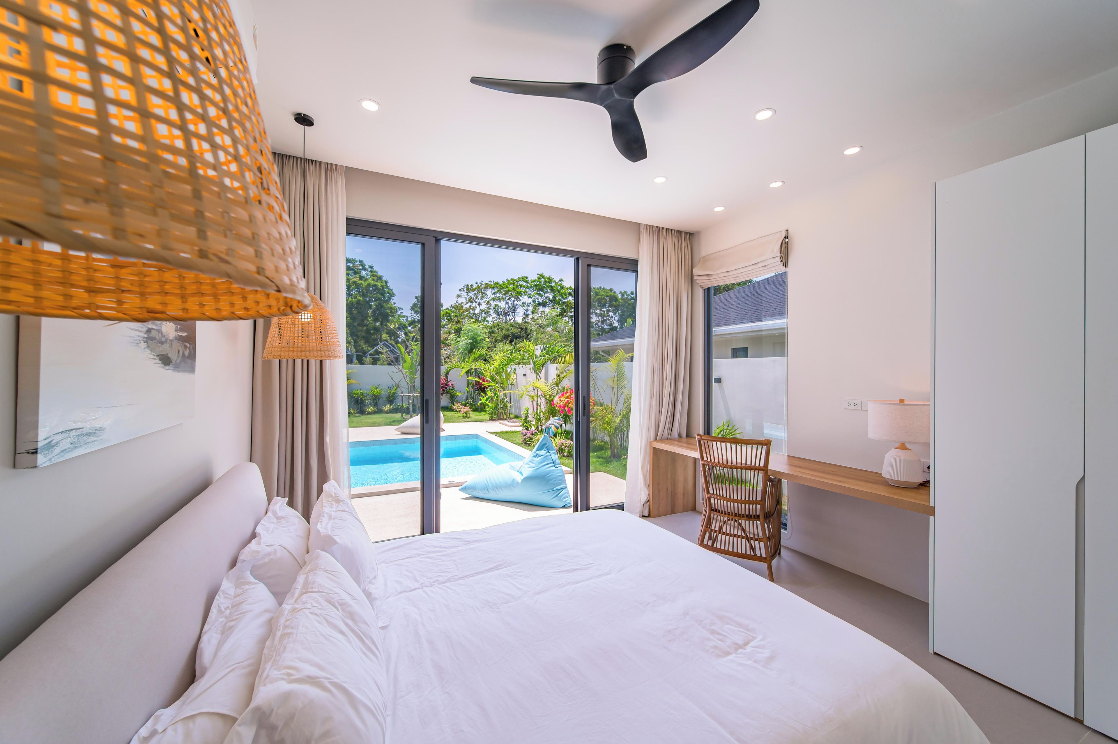 A bedroom with a bed, a desk, a chair, a ceiling fan, and a view of a swimming pool.
