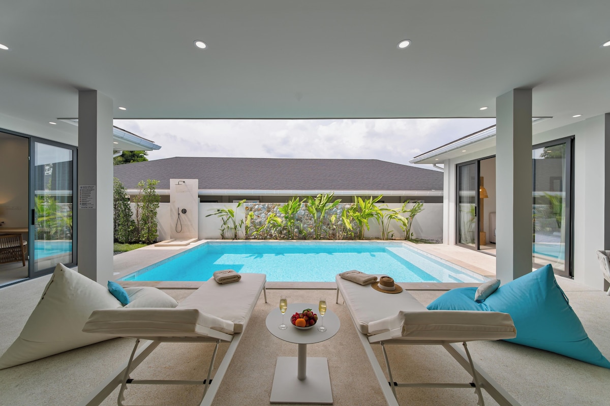 A modern outdoor pool area with lounge chairs, a table with a fruit bowl and glasses, and a view of a house and greenery.