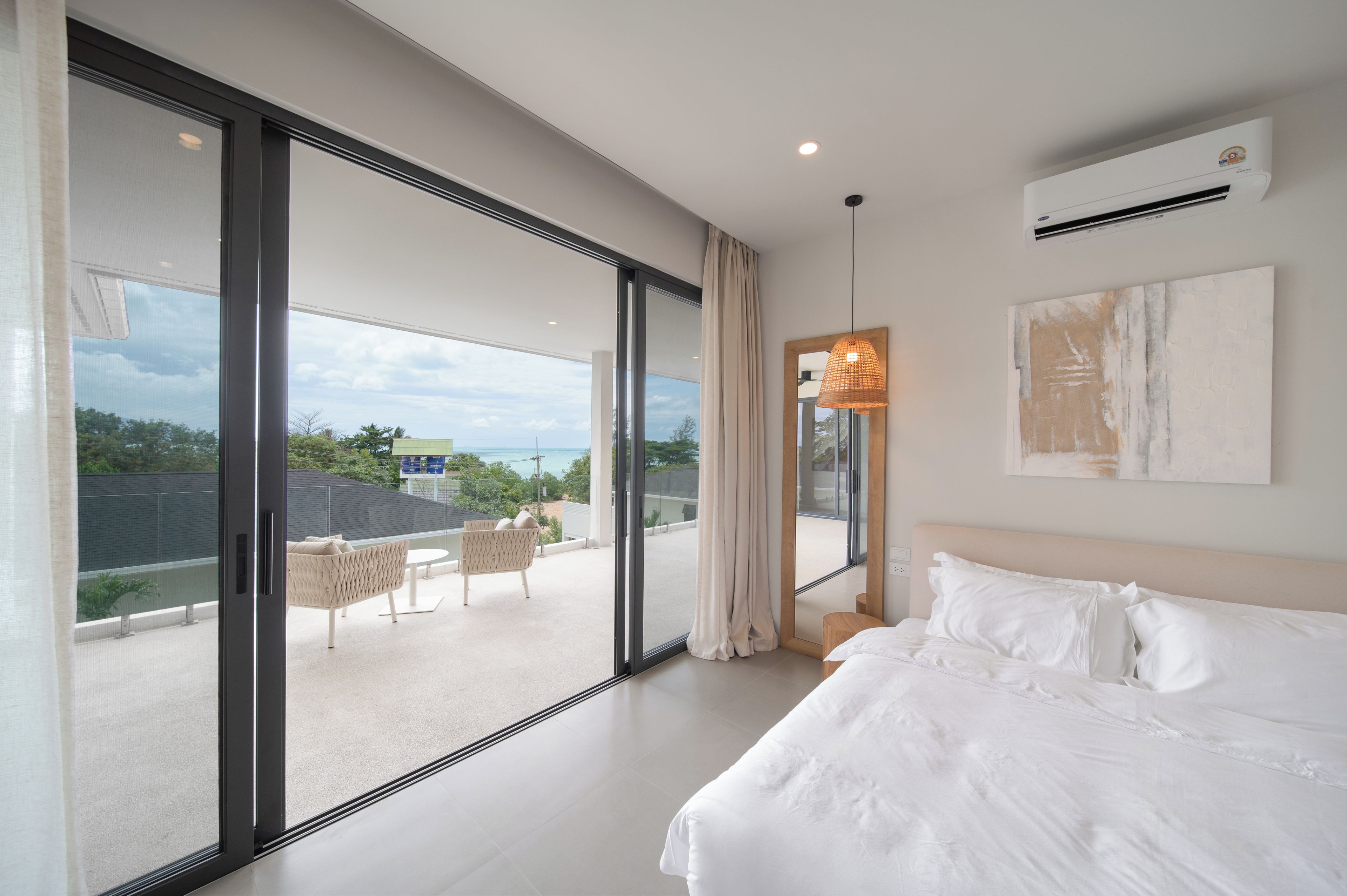 A modern bedroom with a large bed, a view of the outdoors through floor-to-ceiling windows, and a ceiling-mounted air conditioning unit.