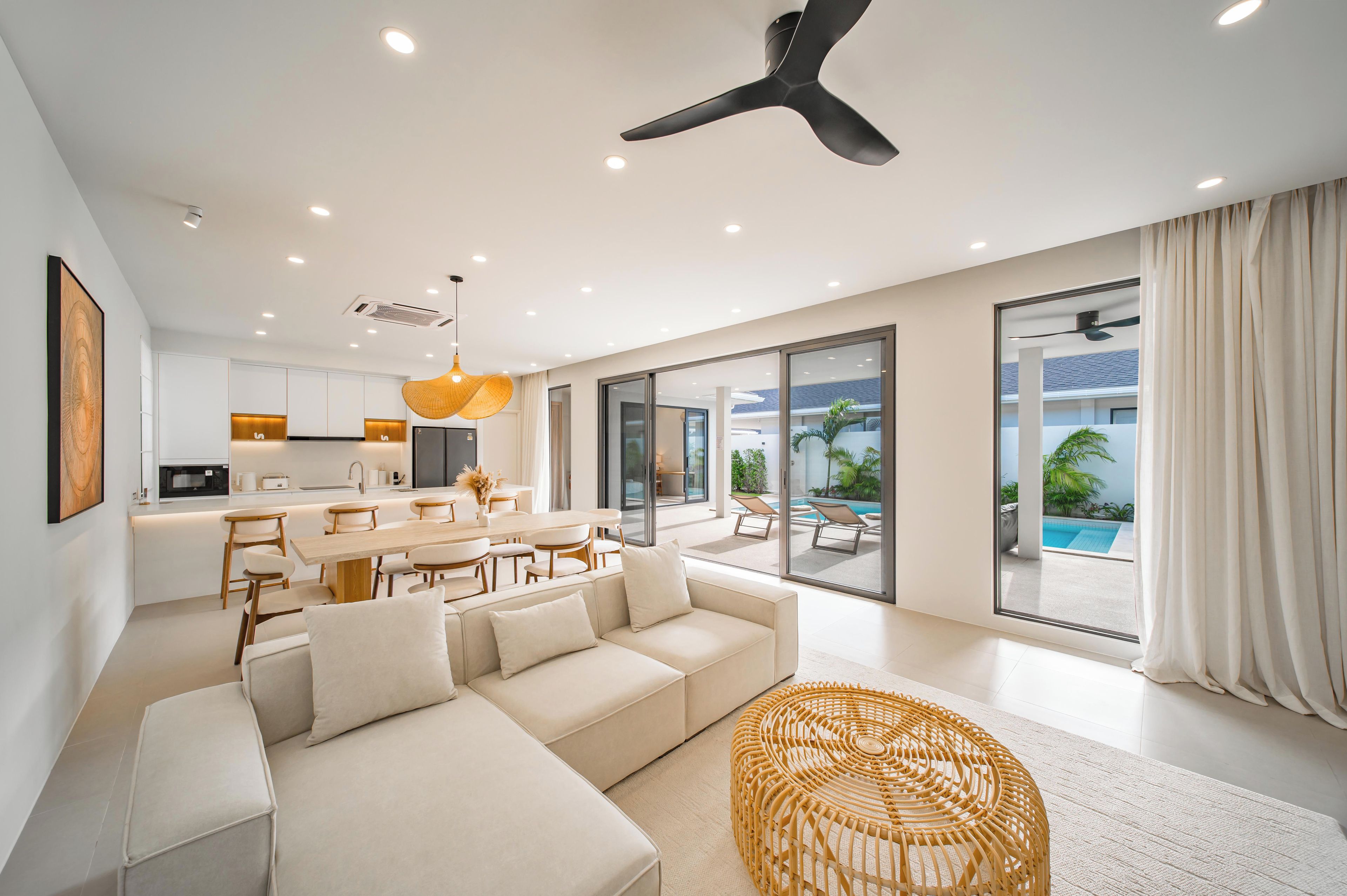 A modern living room with a sofa, dining table, and chairs, a kitchen in the background, and a large sliding glass door leading to a pool area.
