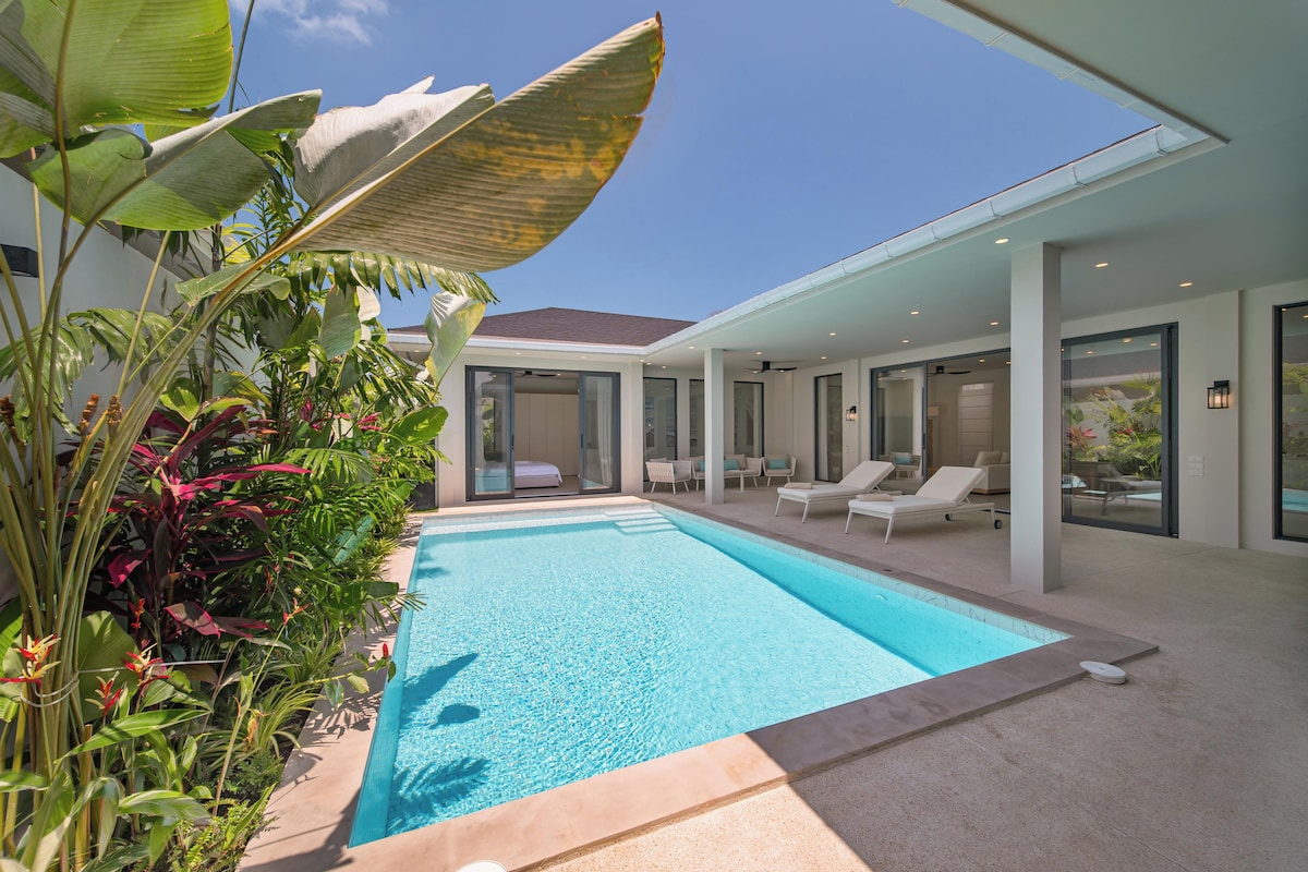 A modern outdoor pool area with a clear blue pool, surrounded by lush tropical plants and a contemporary outdoor lounge with seating.