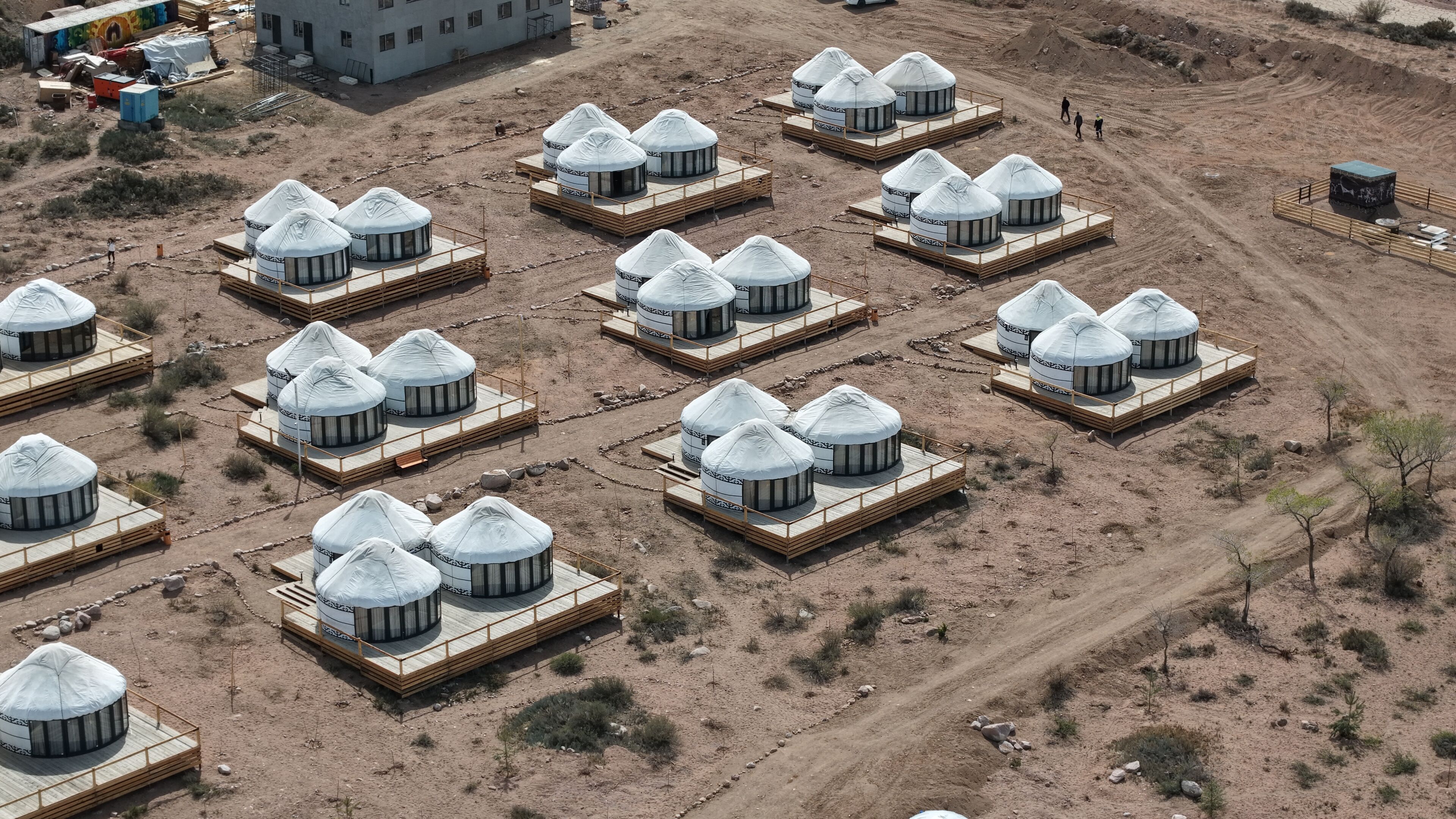 A series of yurts arranged in a grid pattern on a dirt ground with a building in the background.