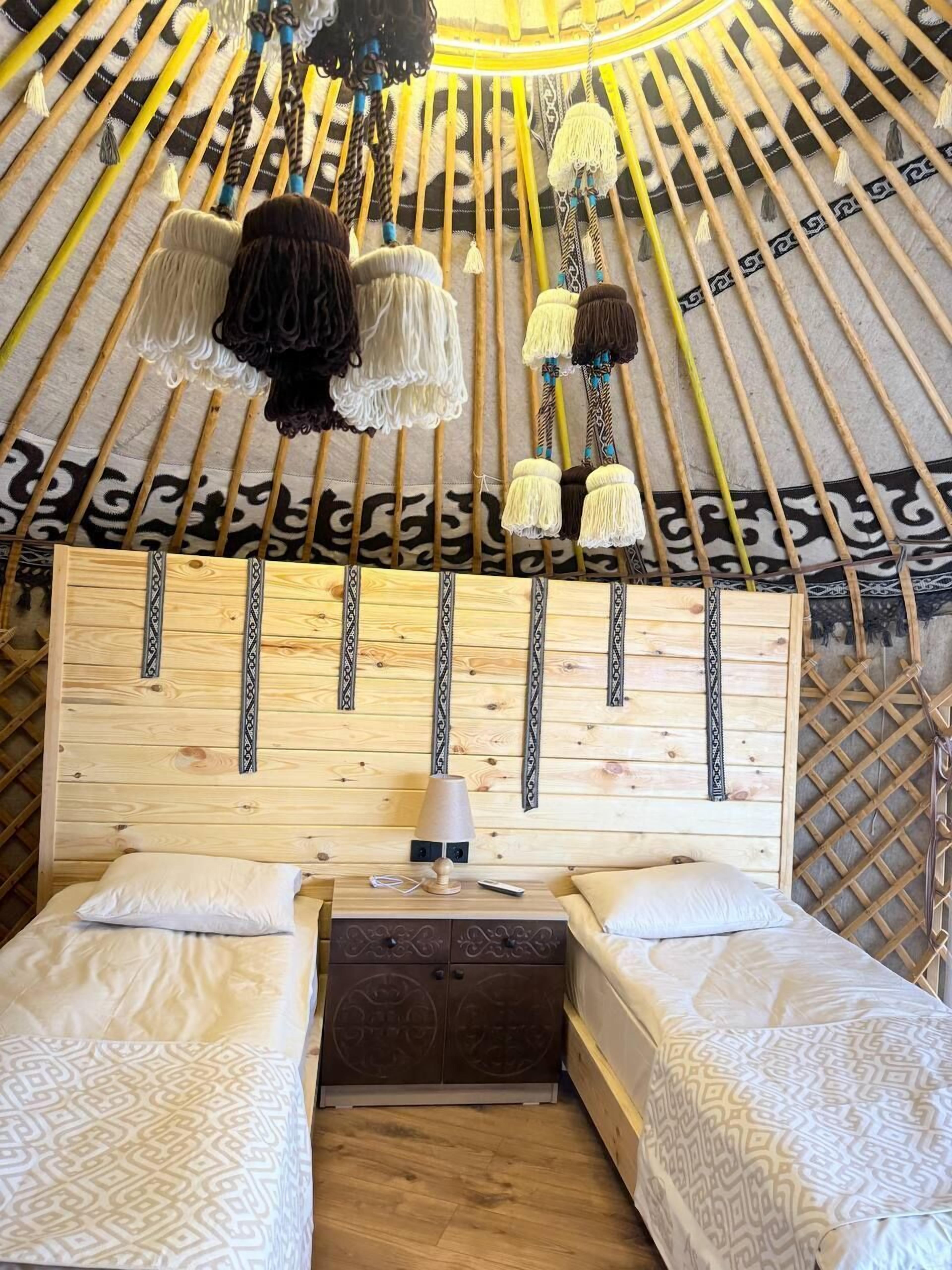 A yurt interior with a wooden headboard, two beds, a nightstand, and hanging decorations.
