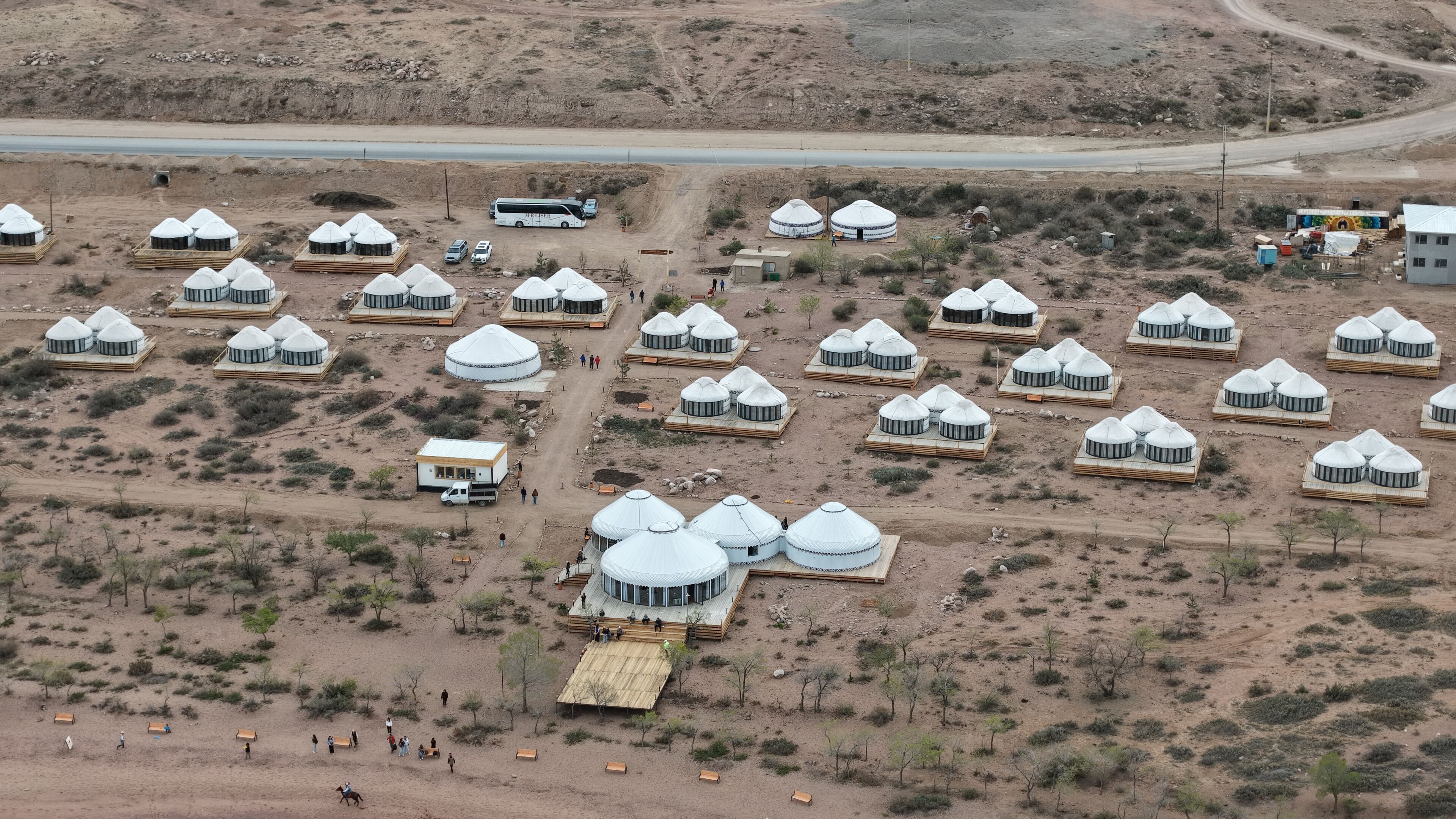 A series of yurts arranged in a semi-circular pattern with a central building, a dirt road, and sparse vegetation.