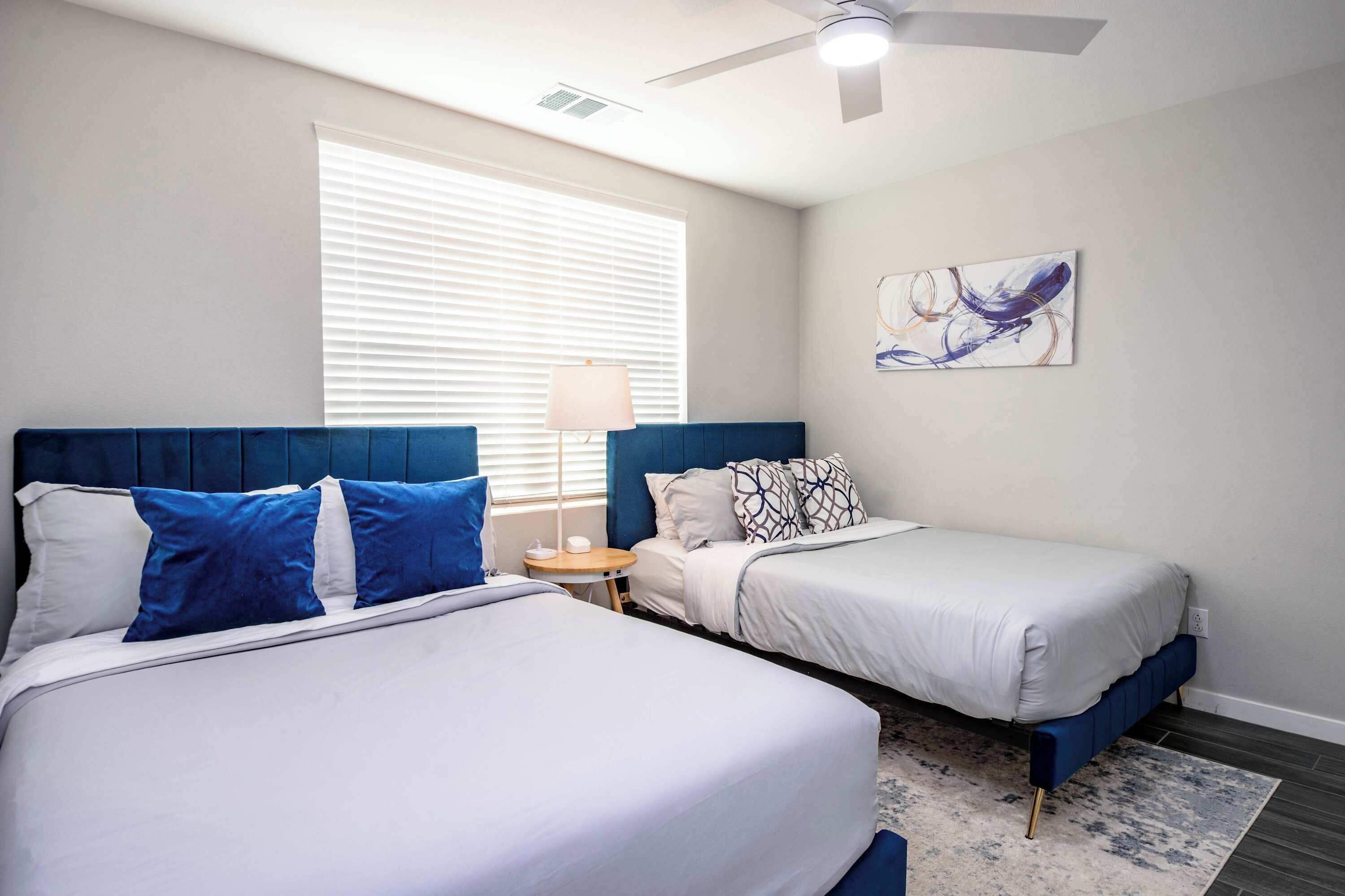 A bedroom with two beds, a ceiling fan, a window with blinds, a small table with a lamp, and a piece of abstract art on the wall.