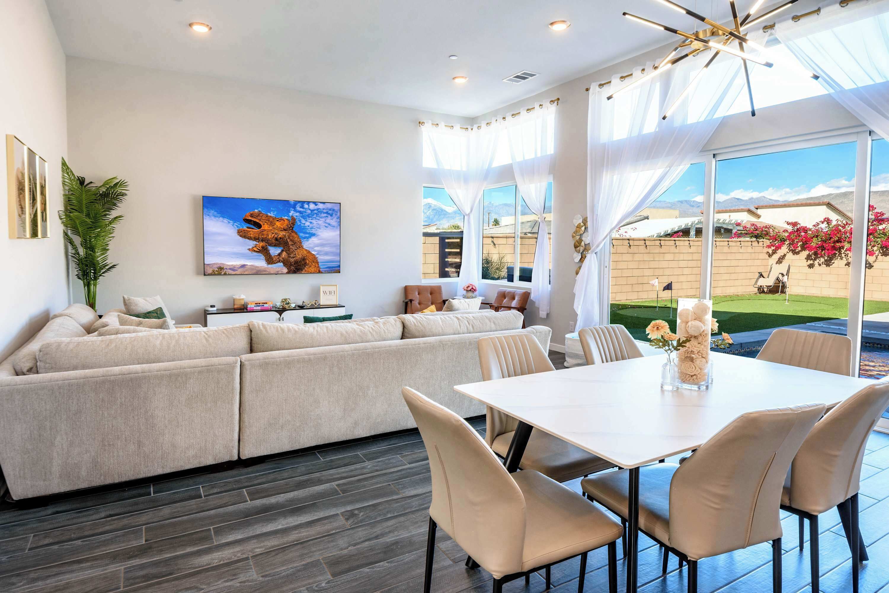 Un salon moderne avec un canapé d’angle, une table à manger avec des chaises, une grande fenêtre avec des rideaux et un tableau accroché au mur.
