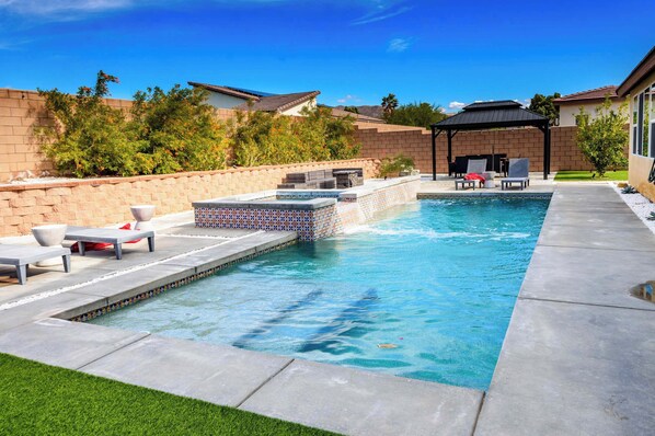 A rectangular swimming pool with a tiled deck, lounge chairs, and a covered patio area.