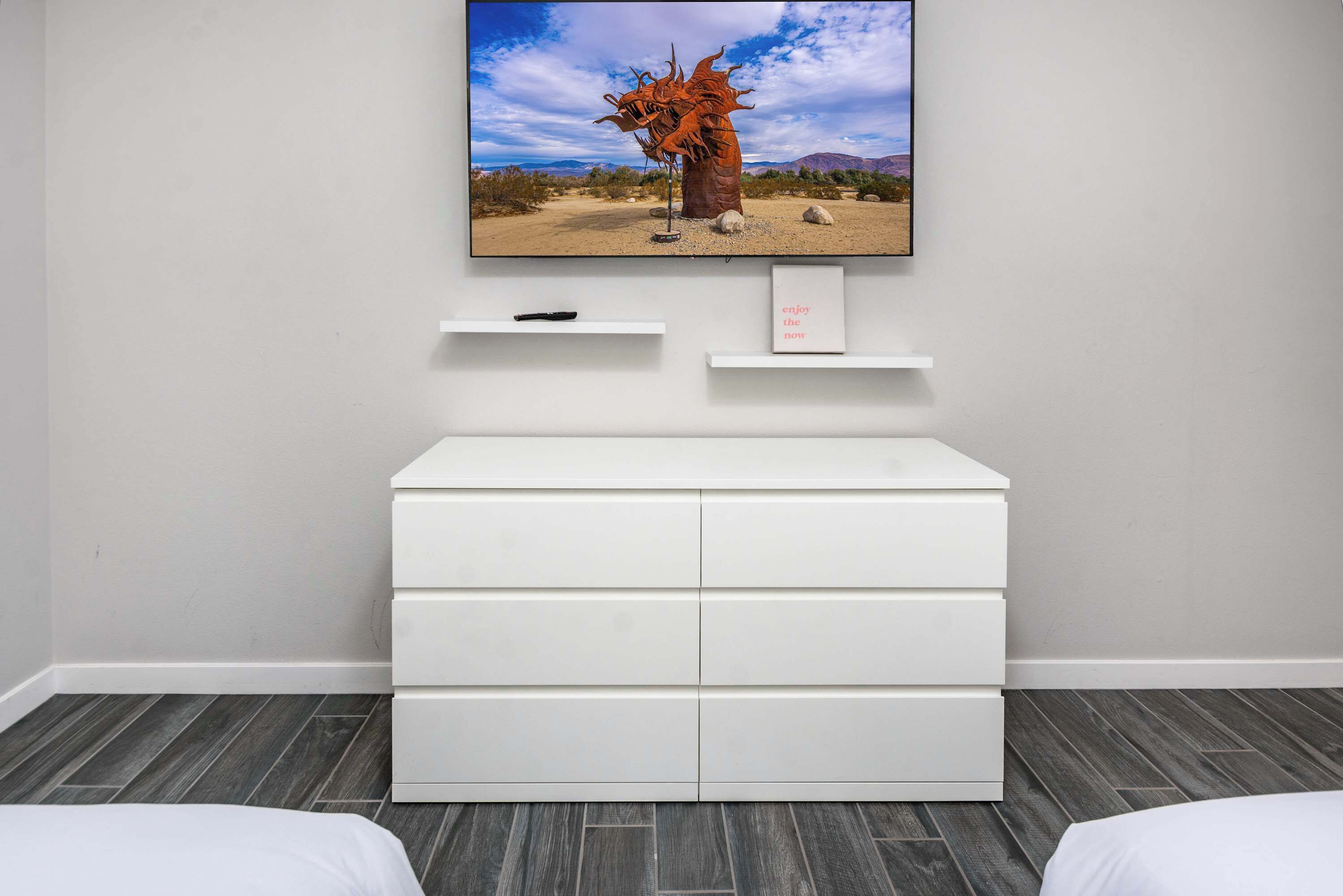 Una camera moderna con una cassettiera bianca, una TV a parete che mostra un paesaggio desertico e un piccolo ripiano con un telecomando.