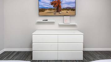 Una camera moderna con una cassettiera bianca, una TV a parete che mostra un paesaggio desertico e un piccolo ripiano con un telecomando.