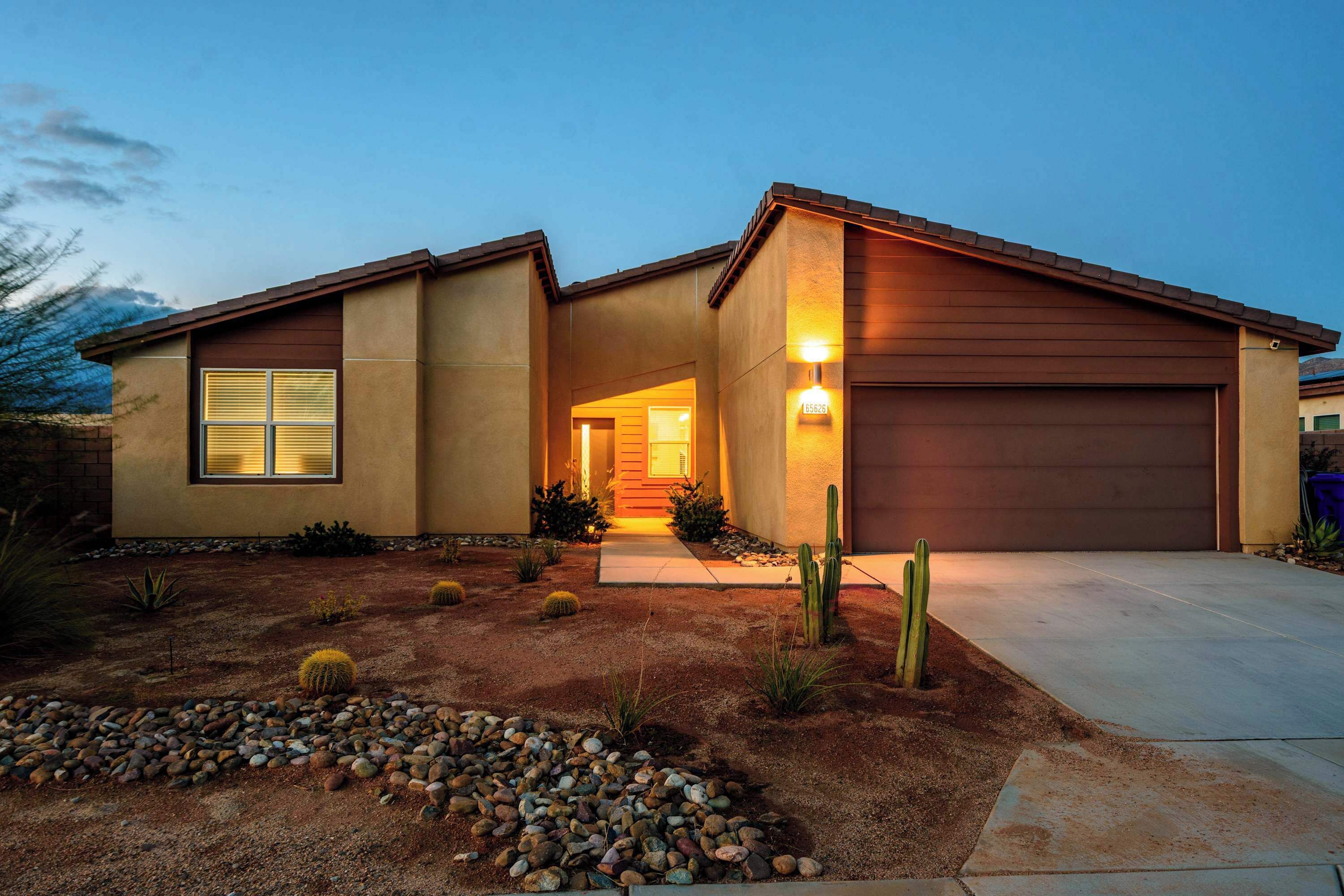 Une maison moderne à deux étages, dotée d’un jardin bien entretenu à l’avant, avec des cactus et un chemin de pierre menant à l’entrée.