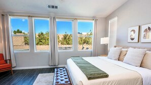 A bedroom with a large bed, a bench, and a view of the outdoors through three windows.