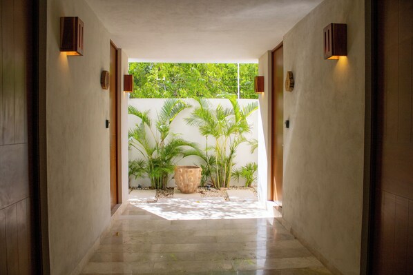 A hallway with a concrete floor, wooden doors, and walls, leading to a garden area with potted plants and a large stone planter. - NHOA 111C Lux 1BRPool access Aldea Zama (Tulum)