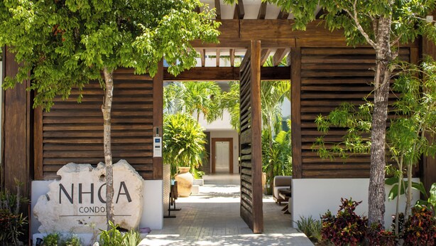 Un ingresso con pergola in legno a una proprietà chiamata NHOA CONDO, con un vialetto lastricato che conduce a una zona salotto e una porta sullo sfondo.