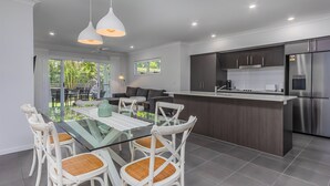 A modern kitchen with stainless steel appliances, a dining area with glass-top table and wicker chairs, and a view of the outdoors. - Dianella Dunes (Casuarina)