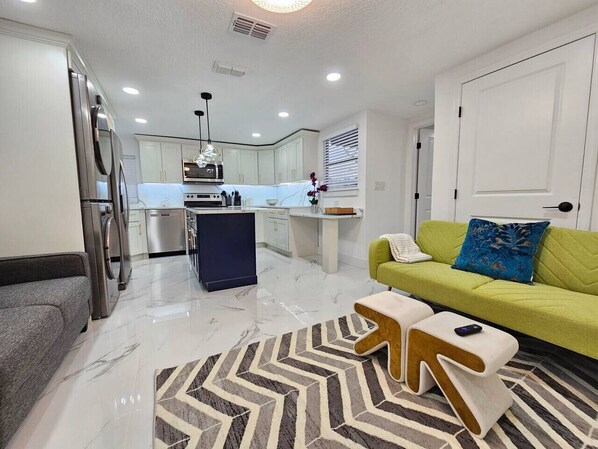 A modern kitchen with white cabinets, a built-in oven, and a central island. There is a green sofa with a blue pillow and a patterned rug on the floor. - Funky Remodeled Otown Bungalow Sleeps 8 (Winter Park)