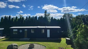 A modern RV-style mobile home with a front porch, surrounded by a well-maintained lawn and dense forest.