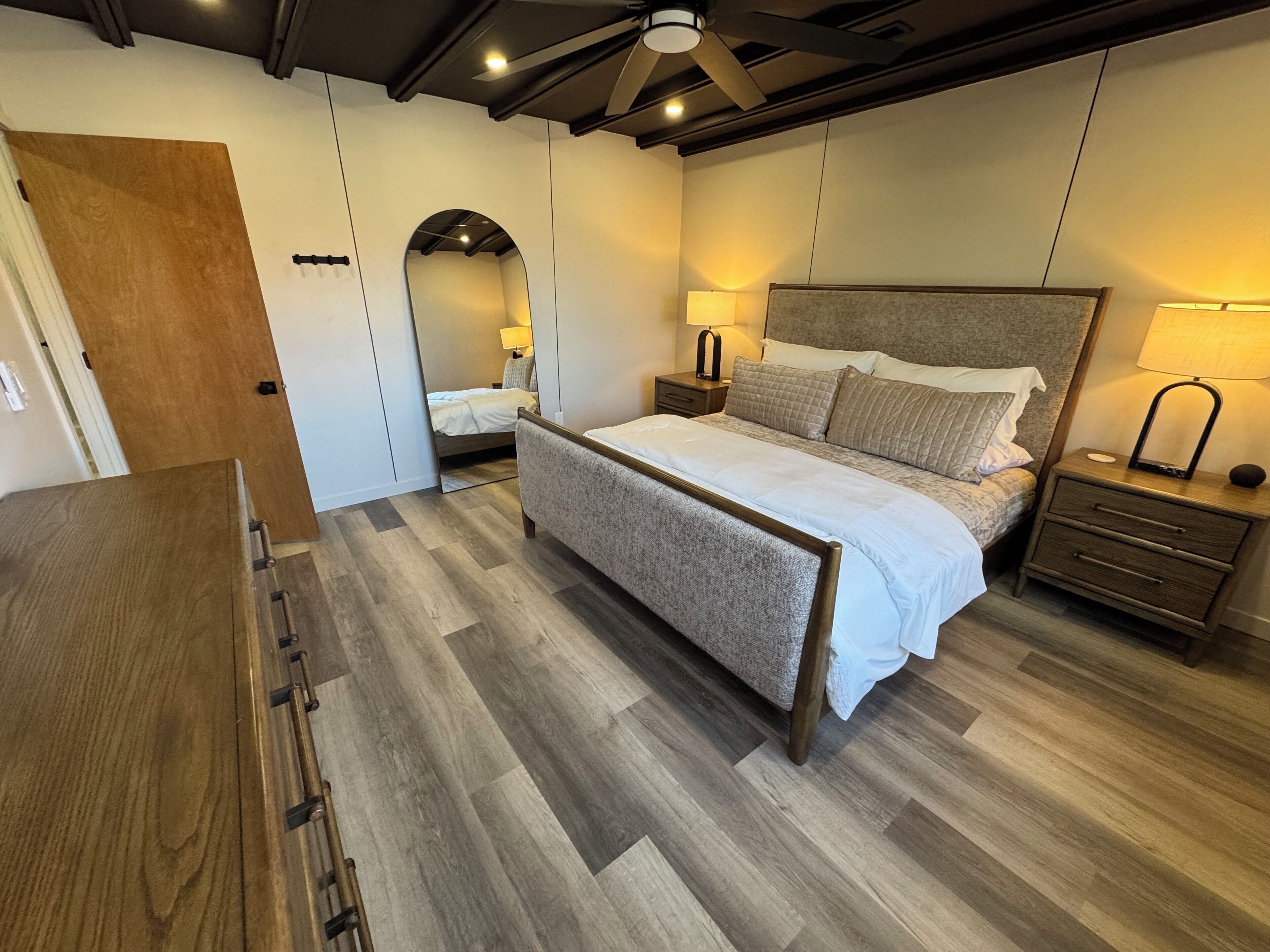 Une chambre avec un plancher en bois, un lit doté d’une tête de lit grise, deux tables de chevet avec des lampes et un miroir au mur.