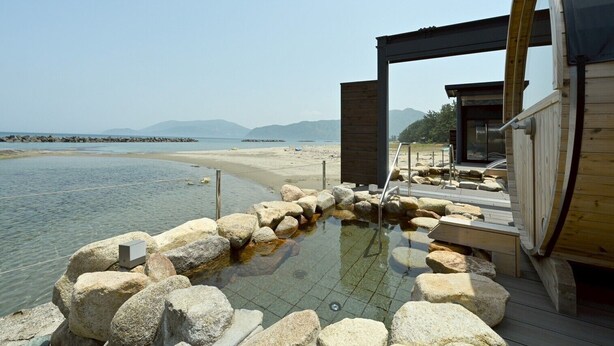 A hot spring bath area with a wooden deck, stone seating, and a view of the beach and mountains. - SIOKUMIHAMA  Sea  Side  Villa  HAMATSUZURI (Miyazu)