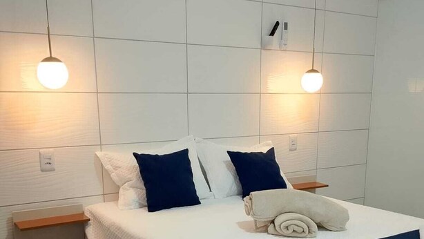 Una cama bien hecha con colcha blanco y azul marino, dos almohadas azul marino y una toalla doblada. Hay dos mesitas de noche con tapas de madera y cajones blancos. La habitación tiene paredes de baldosas blancas y un techo blanco.