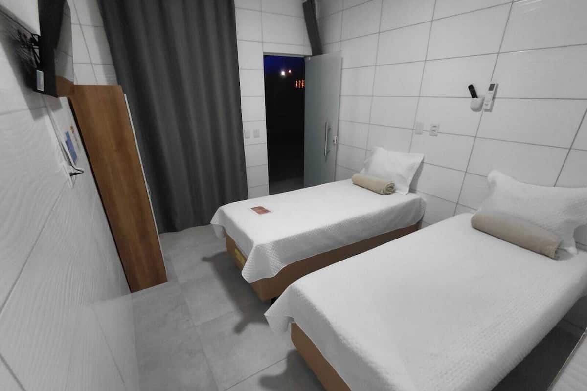 Ein Hotelzimmer mit zwei Betten, einer Holztür, einem grauen Vorhang und einer weiß gefliesten Wand.