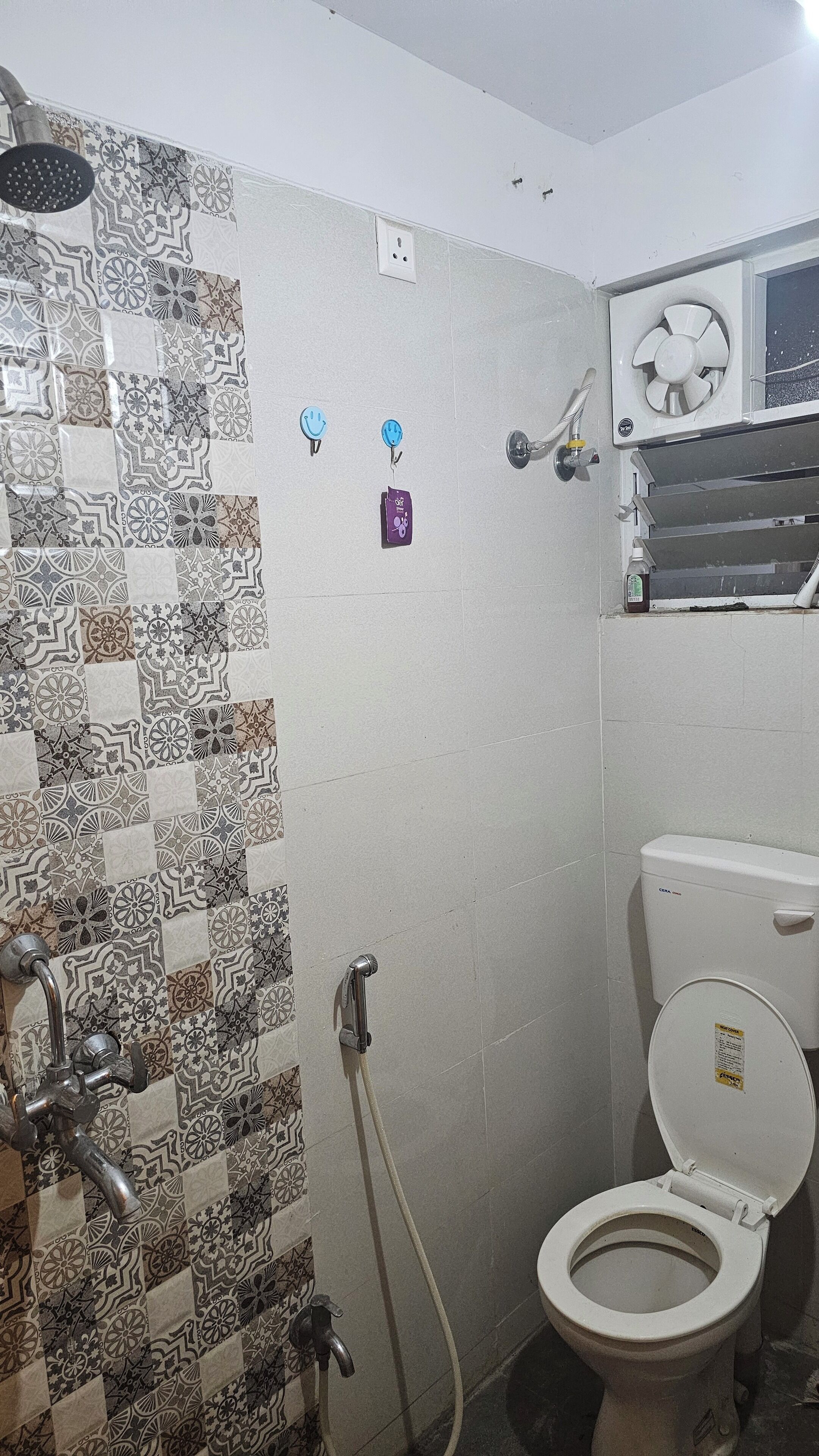 A bathroom with a tiled shower wall, a toilet, and a window with a fan.