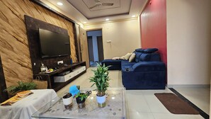 Een moderne woonkamer met een flatscreen televisie, een donkerblauwe bank en een glazen salontafel met potplanten.