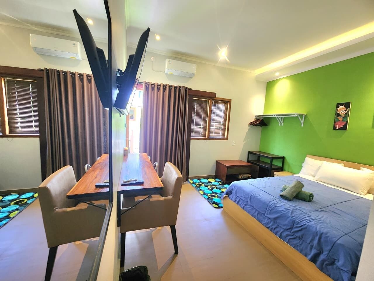 A hotel room with a bed, a desk, and a chair. There is a window with curtains, a green wall, and an air conditioning unit.