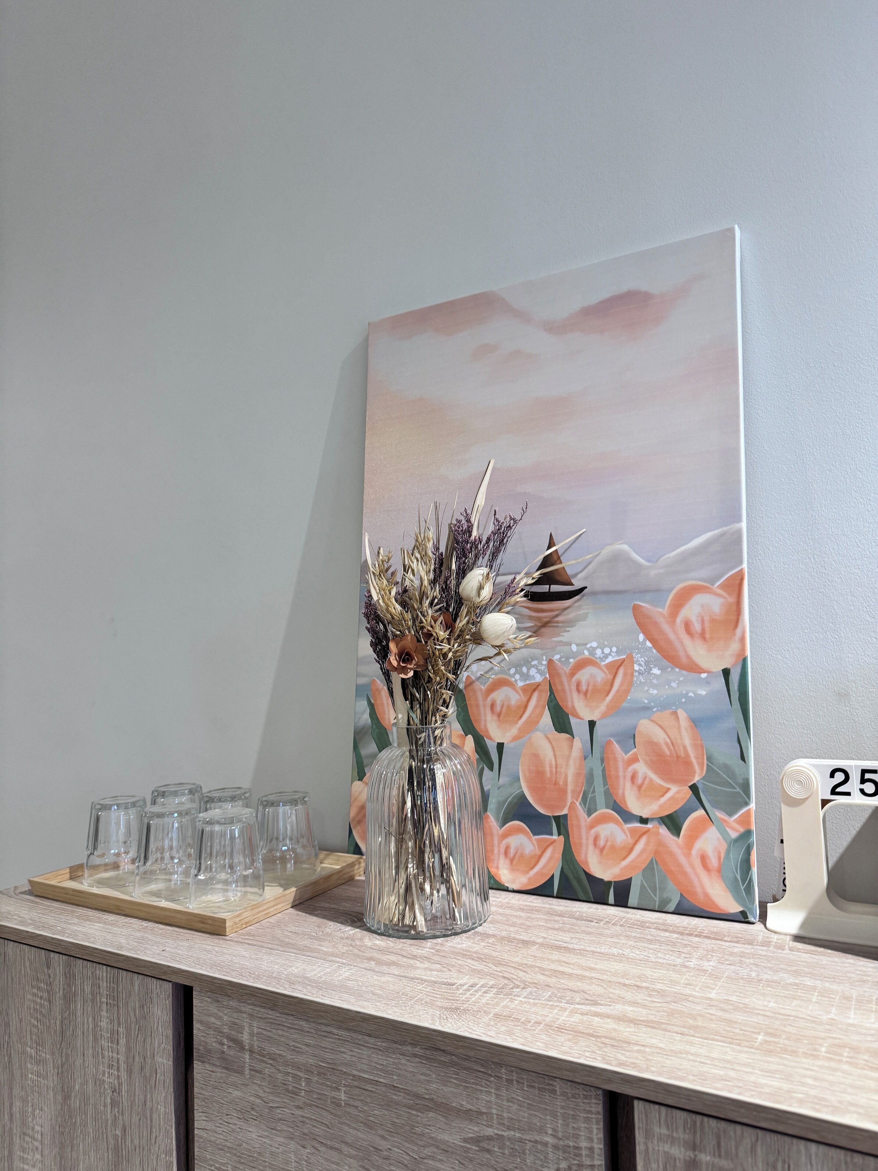 A wooden counter with a glass vase of dried flowers, a framed painting of tulips, and a clock displaying the time.
