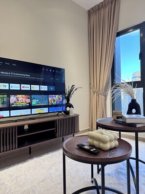Un soggiorno moderno con una TV a schermo piatto che mostra l'interfaccia di un servizio di streaming, un mobile TV in legno, un tavolino rotondo con oggetti decorativi e vista sulla città attraverso un'ampia finestra.