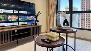 A modern living room with a large flat-screen TV displaying a streaming service interface, a wooden entertainment unit, a round side table with a remote control, and a view of city buildings through a large window. - Comfy Damansara Petaling Jaya 3BR2B  (Petaling Jaya)