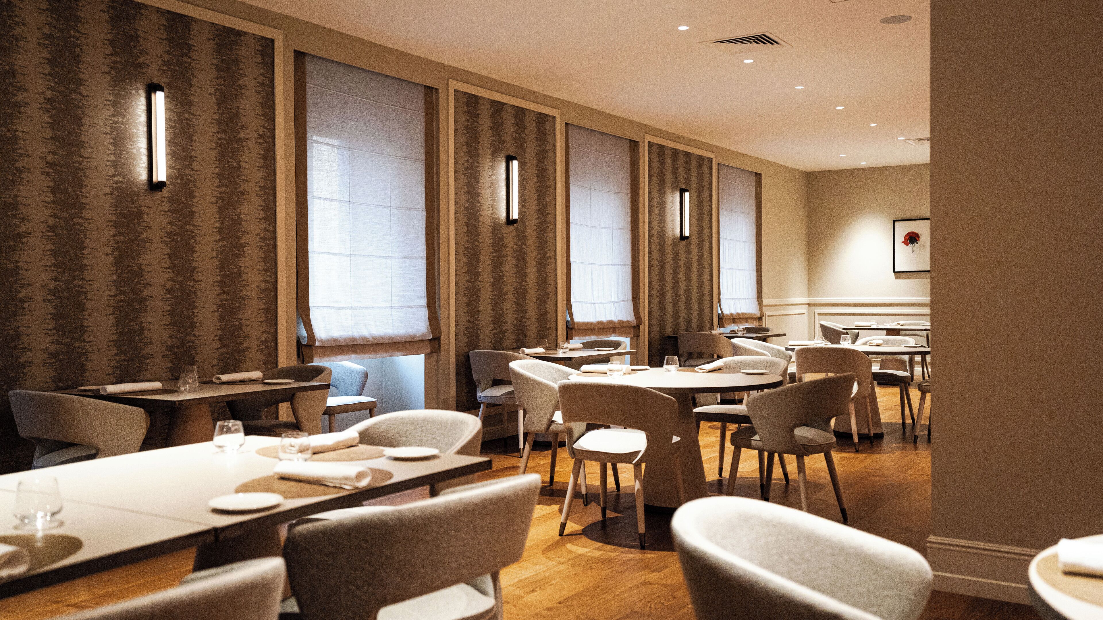 Un comedor de hotel con varias mesas y sillas, amplias ventanas con cortinas y una decoración interior moderna.