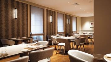 En restaurant i et hotel med flere borde og stole, store vinduer med gardiner, og et moderne indretning.