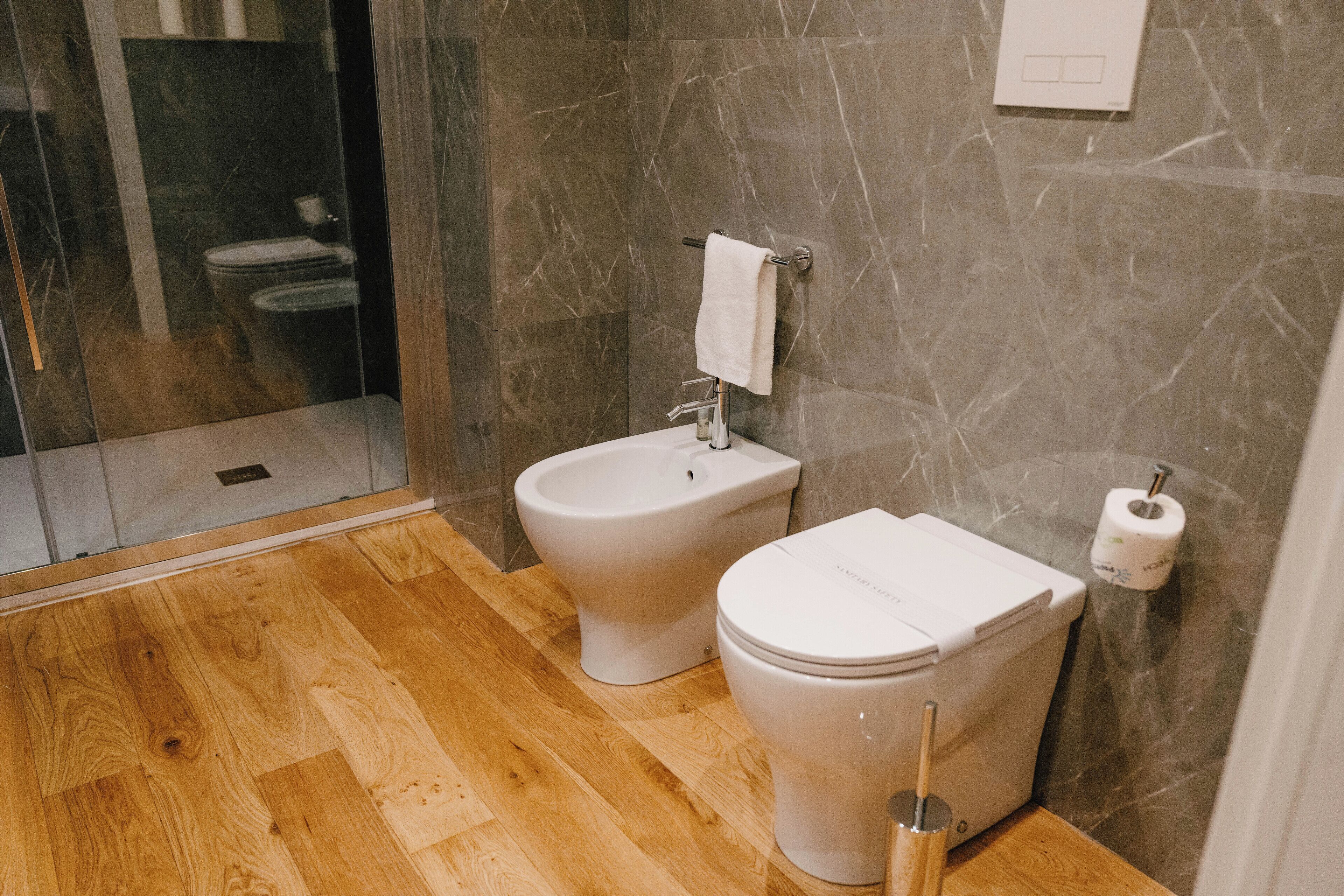 A bathroom with a bidet, toilet, wooden flooring, marble walls, and a glass shower enclosure.