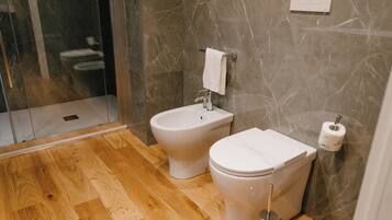 A bathroom with a bidet, toilet, wooden flooring, marble walls, and a glass shower enclosure.