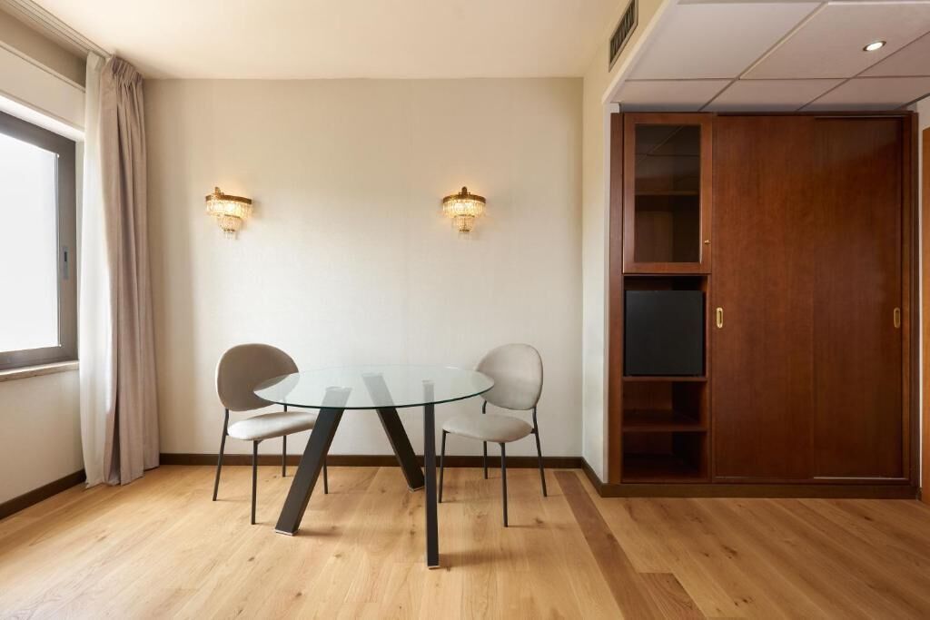 A room with a glass table and two chairs, a wooden wardrobe, and two wall-mounted lights.
