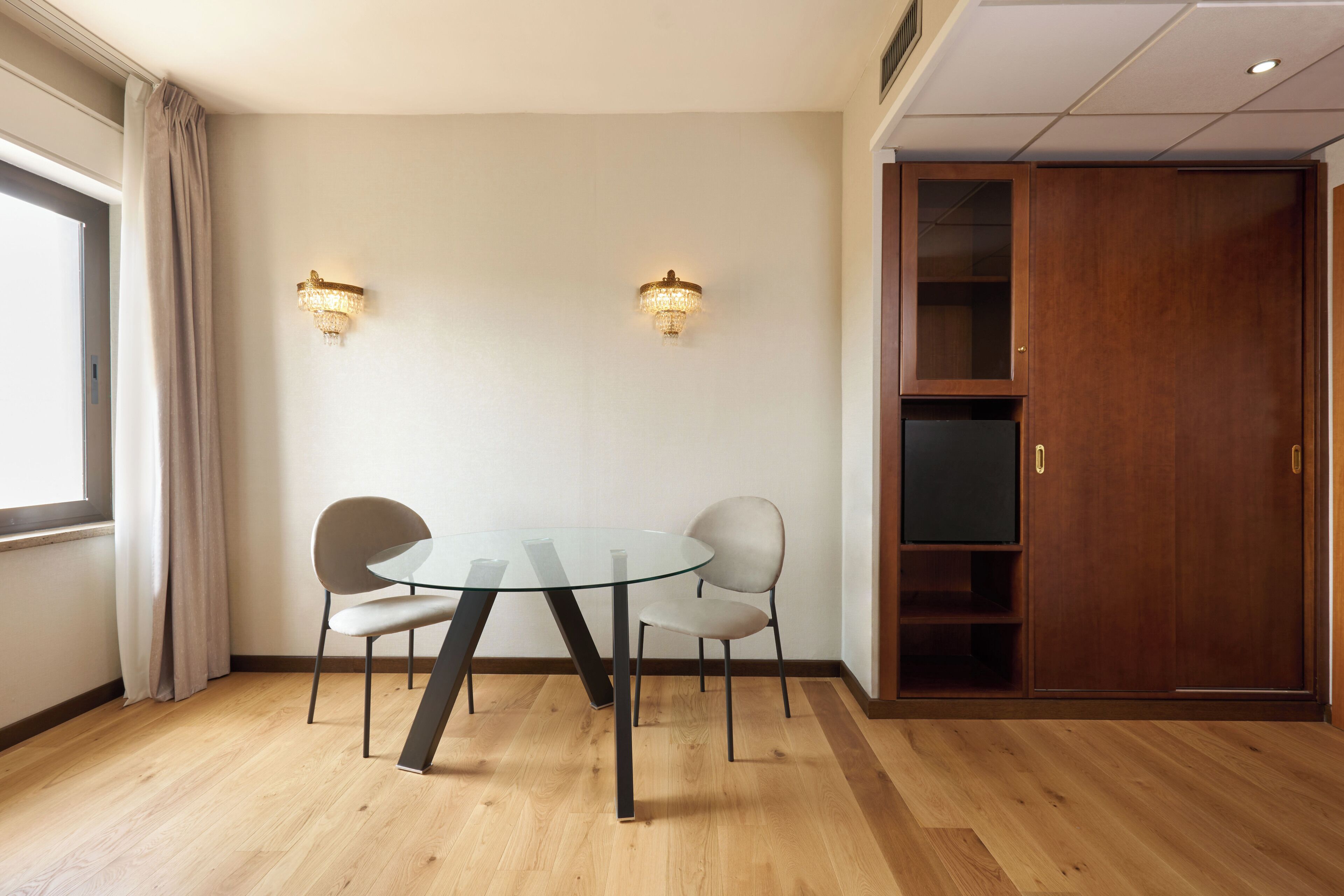 Une pièce avec une table en verre et quatre chaises, un plancher en bois, une armoire en bois et deux lampes fixées au mur.