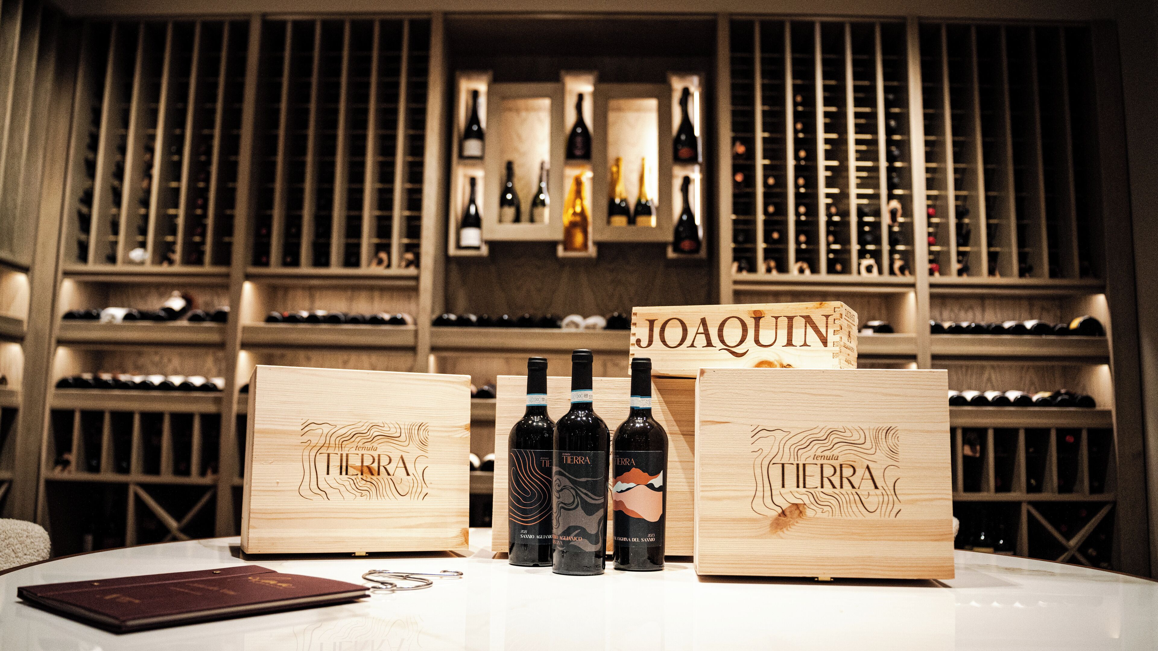 Wooden wine boxes with bottles labeled 'TIERRA' and 'JOAQUIN' in a wine cellar with shelves of wine bottles.