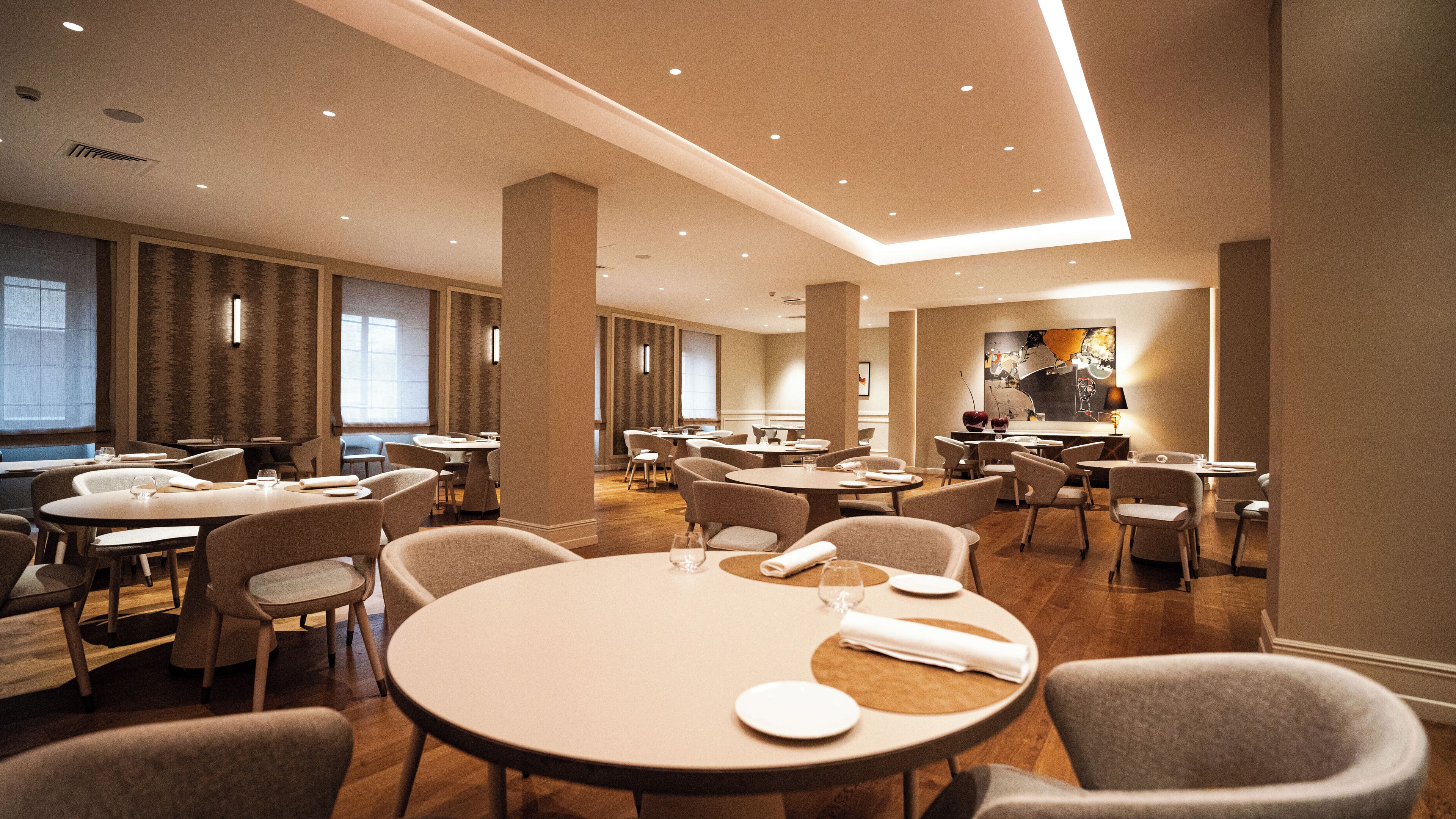Een ruime hotel eetruimte met meerdere ronde tafels en stoelen, grote ramen en een modern plafondontwerp.
