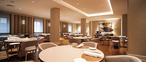 Een ruime hotel eetruimte met meerdere ronde tafels en stoelen, grote ramen en een modern plafondontwerp.