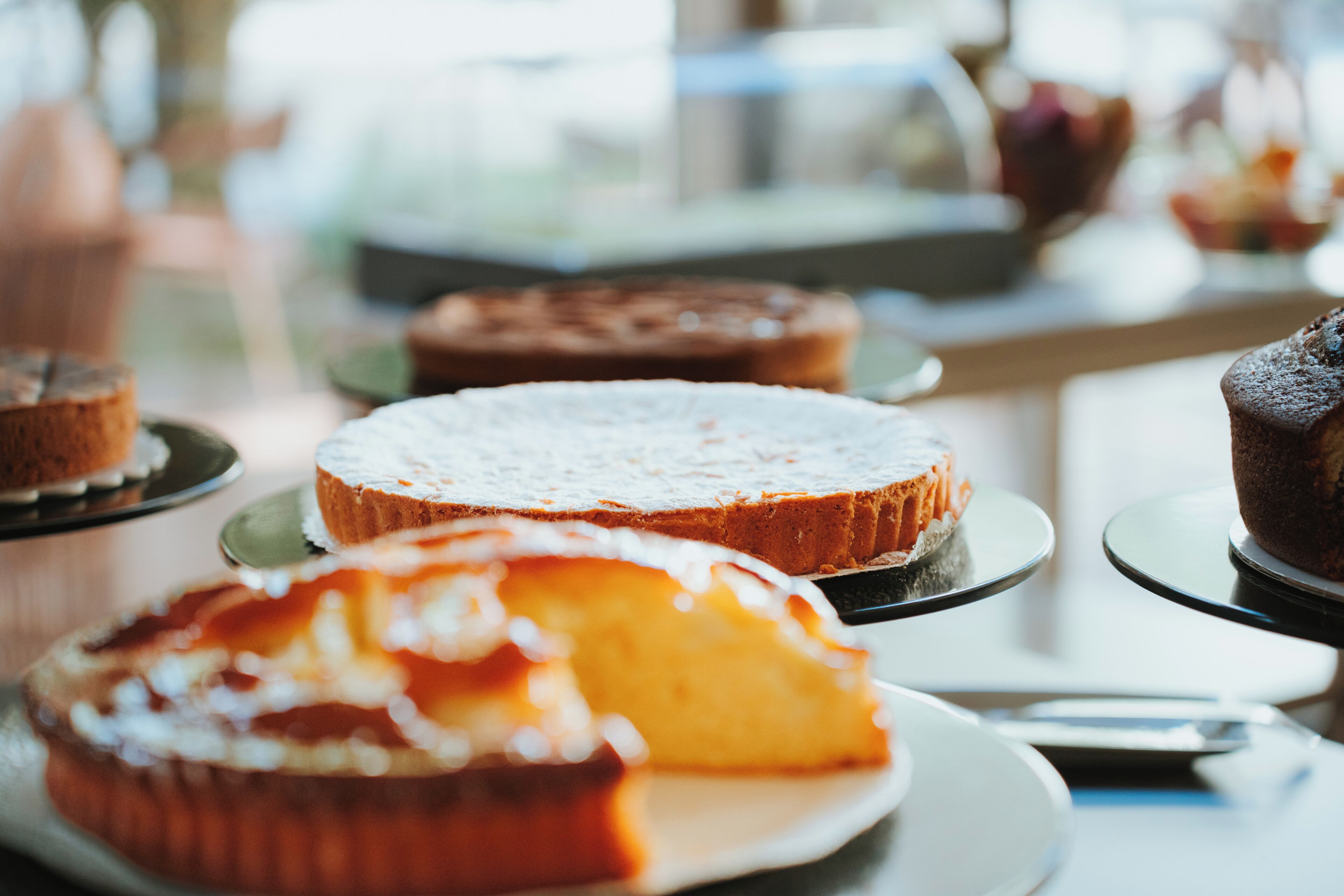 A variety of cakes on display, including a slice of peach cake with a dusting of powdered sugar.