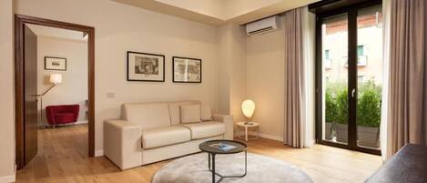 Una sala moderna con un sofá color beige, una mesa de centro redonda y un sillón rojo. En la pared, dos cuadros enmarcados, una mesita de noche con una lámpara, y un ventanal con cortinas.