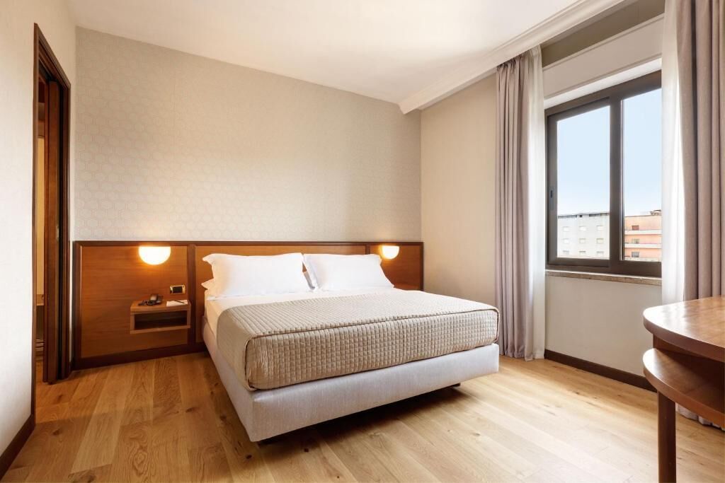 A hotel room with a bed, bedside table, and a window with curtains.