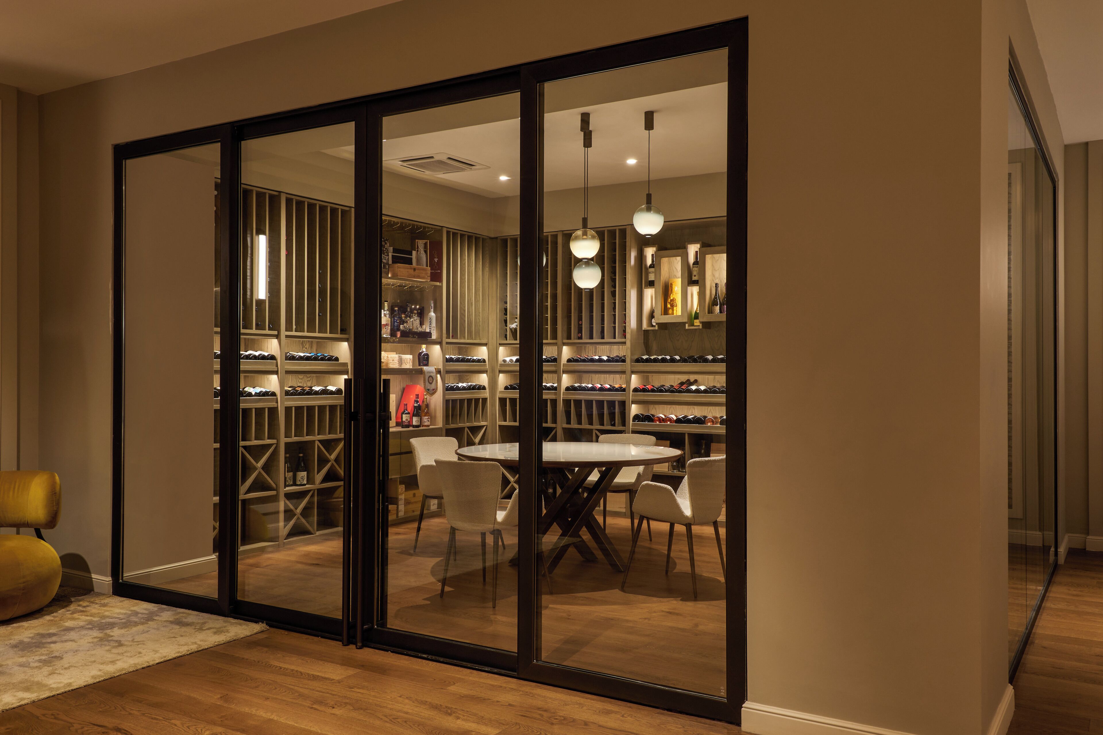Una habitación moderna con puerta de vidrio corredera que da a una bodega de vinos, una mesa de comedor redonda con sillas e iluminación colgante.
