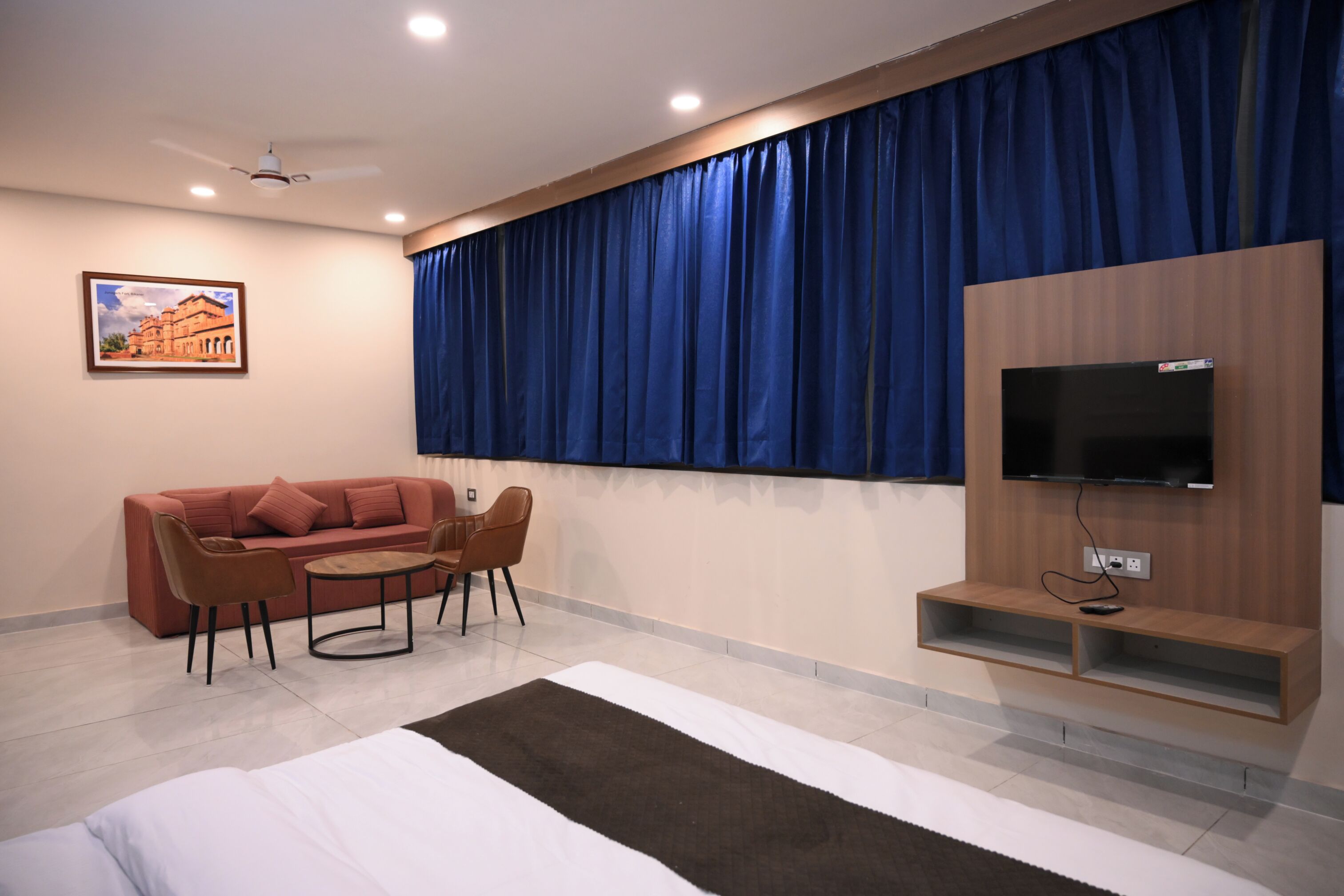 A hotel room with a bed, a TV mounted on the wall, a sofa, two chairs, a small table, and a large window with blue curtains.